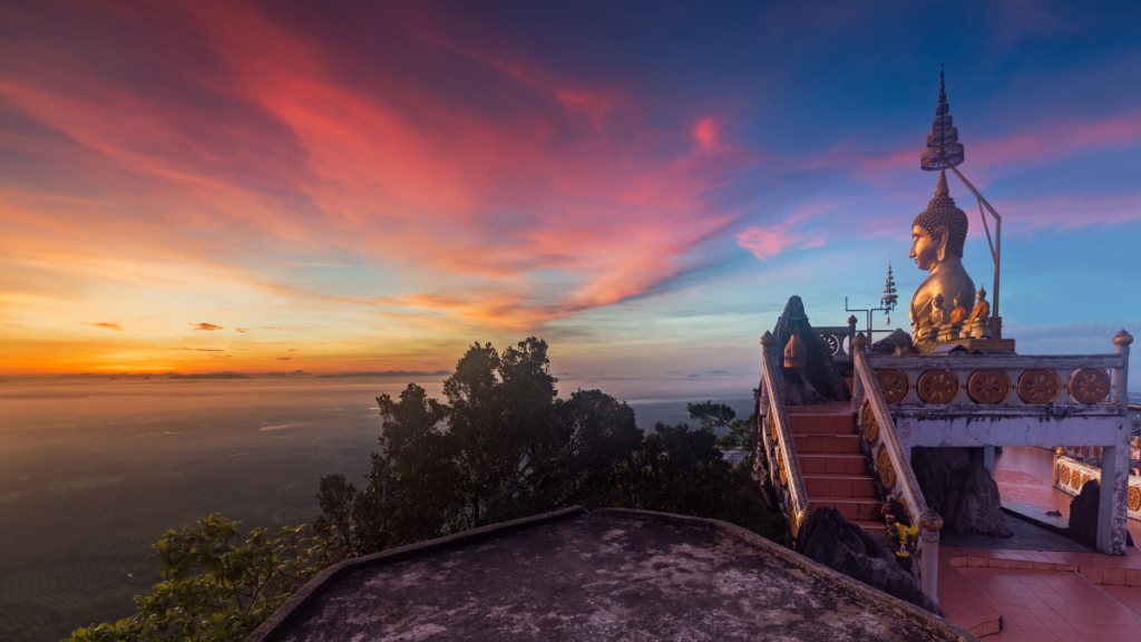 A golden buddha statue seems to contemplate a rainbow-sherbet sunset with wispy clouds as seen from Tiger Cave Temple, Krabi